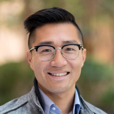 A Chinese-American male smiling with short black hair and glasses, wearing a blue button-up shirt and gray jacket.