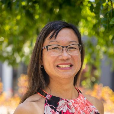 Headshot of Dana Kawaoka-Chen, an Asian-American woman with medium-length black hair and bangs. She is wearing glasses, and is pictured in a floral, pink and black halter top, standing outdoors.