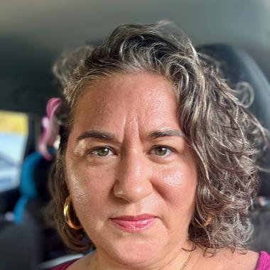 Close-up of Jessamyn Sabbag, an Arab-Anglo woman with tan skin and wavy brown and grey hair, wearing a fuchsia tank top and lipstick, and gold jewelry.