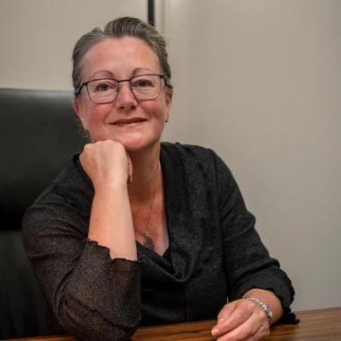 . Middle aged white woman in dark grey sweater and wearing glasses resting her chin on her hand and smiling at camera.