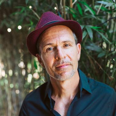 Picture of Mateo Nube from the waist up, wearing a black button down shirt and a cherry colored hat.