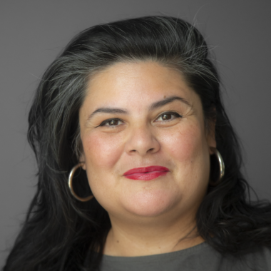 Headshot of woman with long black hair, light brown skin, and red lipstick wearing a gray shirt and standing in front of a gray background.