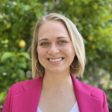 A white woman in her 30s with medium-length blond hair wearing a pink blazer